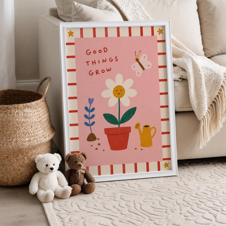 A framed poster with a pink background and illustrations of a flower, a watering can, and a butterfly, along with the text "Good things grow". Two teddy bears are sitting next to the poster on a rug.