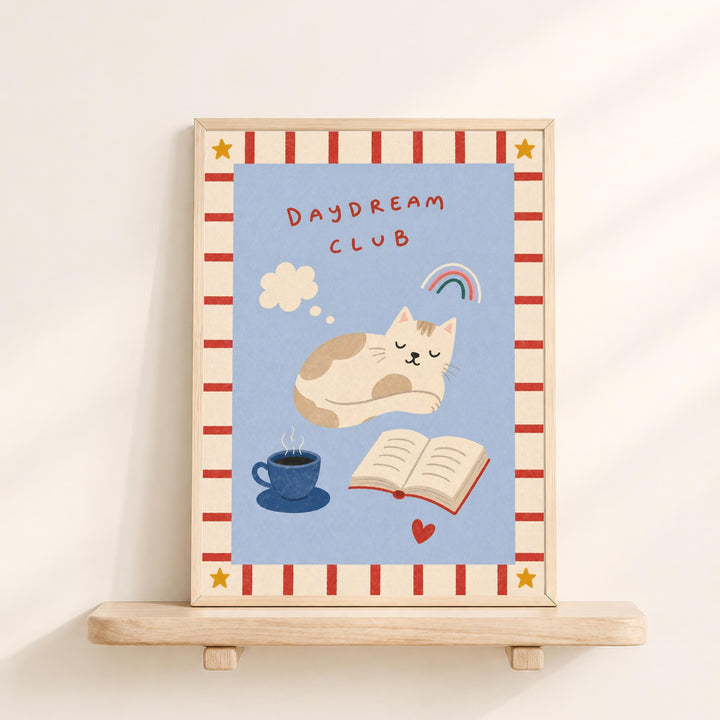 The image is a framed illustration of a cat sleeping on a blue background, with a cup of coffee and a book nearby. The text "DAYDREAM CLUB" is written above the cat, and there is a rainbow and a heart in the background.