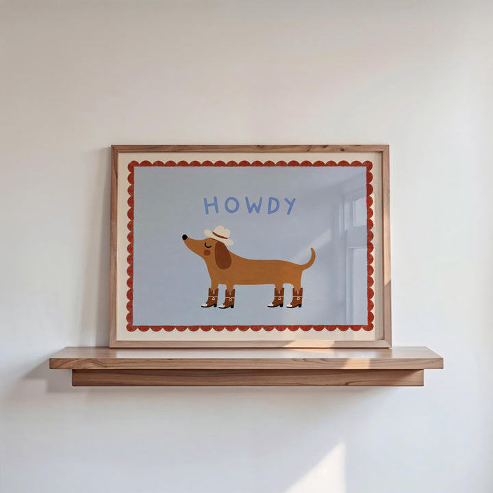 A framed illustration of a dog wearing a cowboy hat, with the word "HOWDY" written above it, is displayed on a wooden shelf against a white wall.