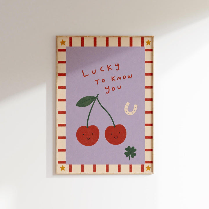 The image is a framed illustration featuring two cherries with smiling faces, a four-leaf clover, and a horseshoe, all set against a purple background with a striped border.