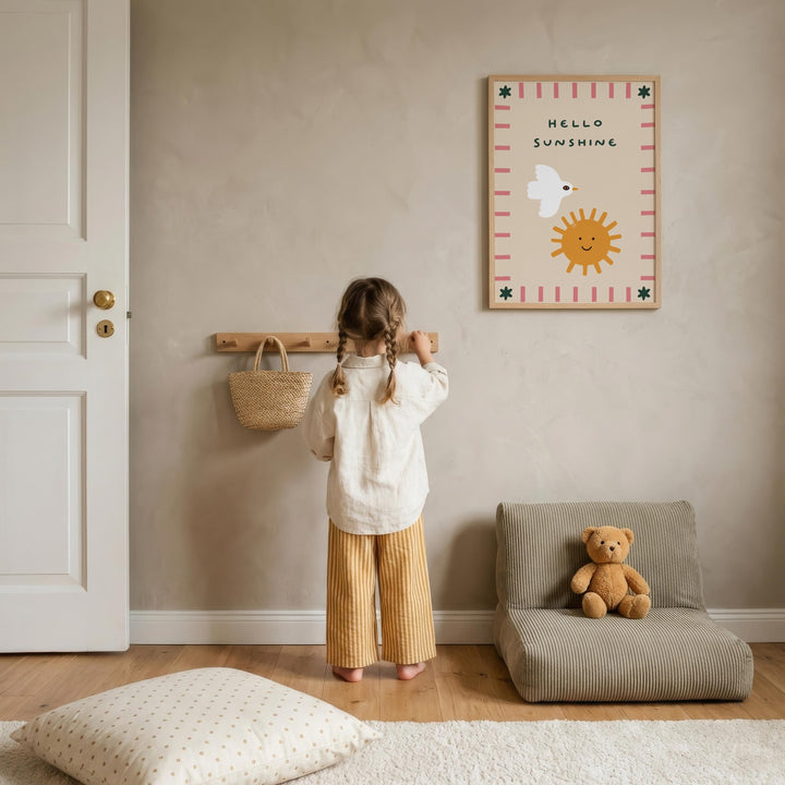 A young girl stands in a cozy room, looking at a framed poster with a cheerful message. The room has a wooden floor, a beige wall, and a small chair with a teddy bear on it.