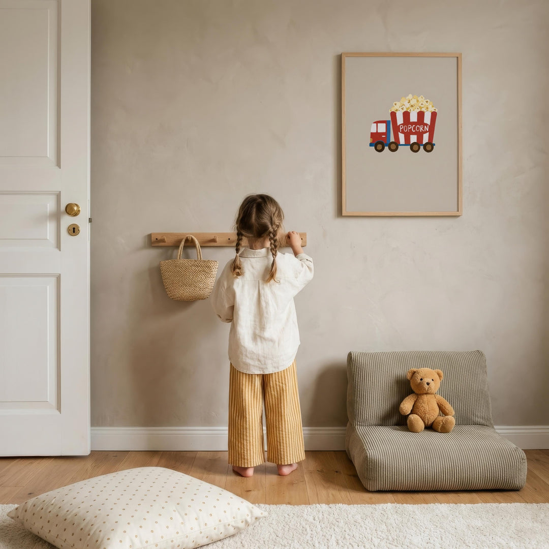A young girl stands in a cozy living room, looking at a framed picture of a red and white popcorn truck on the wall. The room has a beige color scheme, with a teddy bear sitting on a chair nearby.
