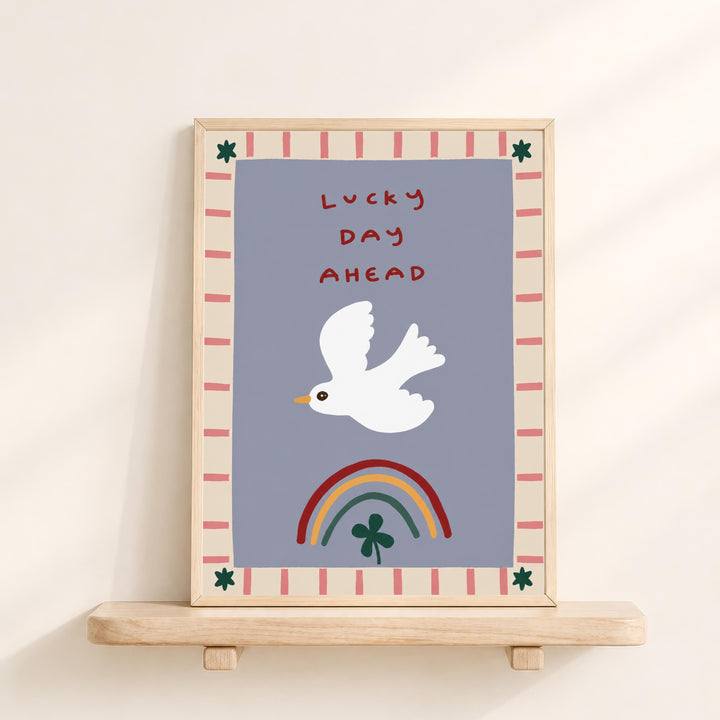 The image is a framed poster with a white bird flying over a rainbow and a clover. The poster has a border with red and pink stripes and the text "Lucky day ahead" written in red.