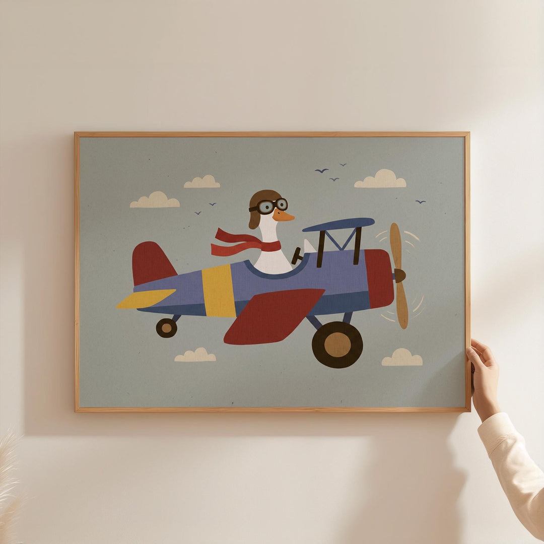 A person is holding up a framed picture of a cartoon goose wearing a pilot's hat and scarf, flying a colorful airplane in the sky.