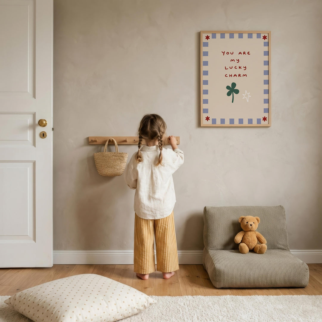 A young girl stands in a cozy room, looking up at a framed sign that reads "You are my lucky charm". The room has a beige color scheme, with a teddy bear sitting on a chair and a wooden shelf with a woven basket nearby.