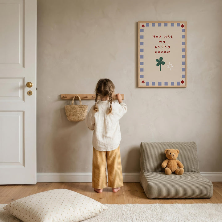 A young girl stands in a cozy room, looking up at a framed sign that reads "You are my lucky charm". The room has a beige color scheme, with a teddy bear sitting on a chair and a wooden shelf with a woven basket nearby.
