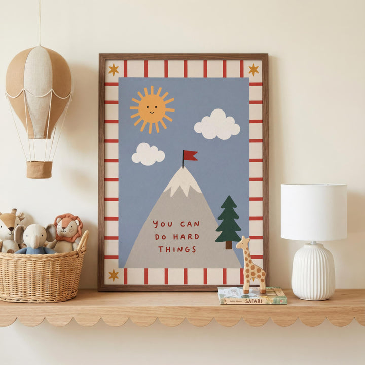 a framed poster with a mountain, a sun, clouds, and a flag, along with a motivational message. The poster is displayed on a shelf, accompanied by a basket of stuffed animals, a lamp, and a small toy giraffe.