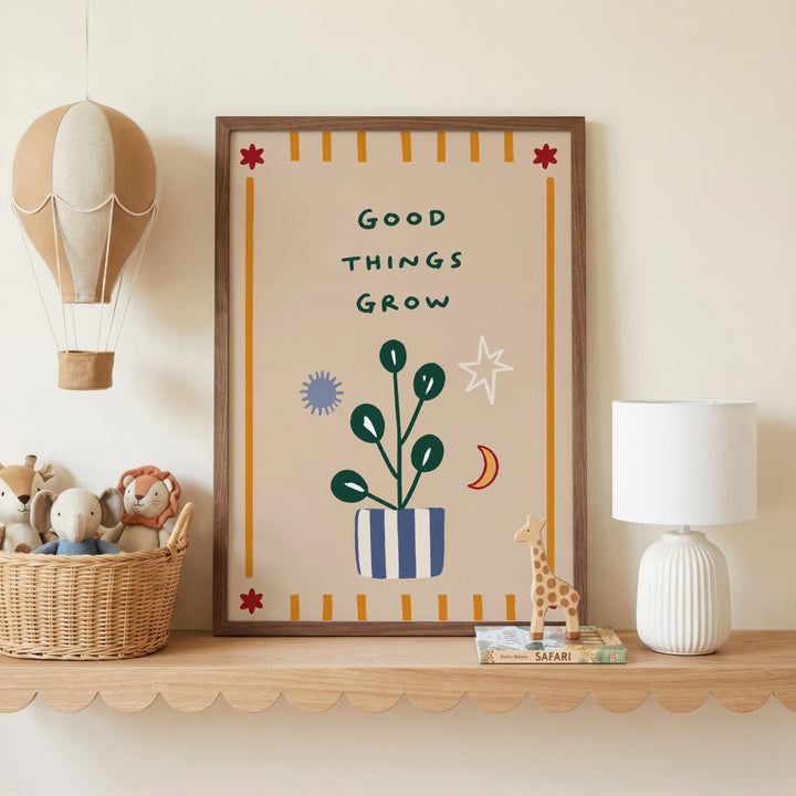 a framed poster with the text "GOOD THINGS GROW" on it, surrounded by various decorative elements such as a hot air balloon, a plant, and a giraffe figurine. The poster is displayed on a shelf, with a basket of stuffed animals and a lamp nearby.