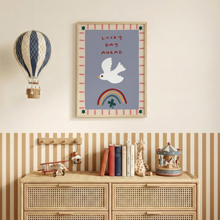 a wall with a framed poster of a white bird, a hot air balloon, and a rainbow, along with the text "Lucky day ahead". Below the poster, there is a wooden dresser with a toy airplane, a carousel, and a collection of books.