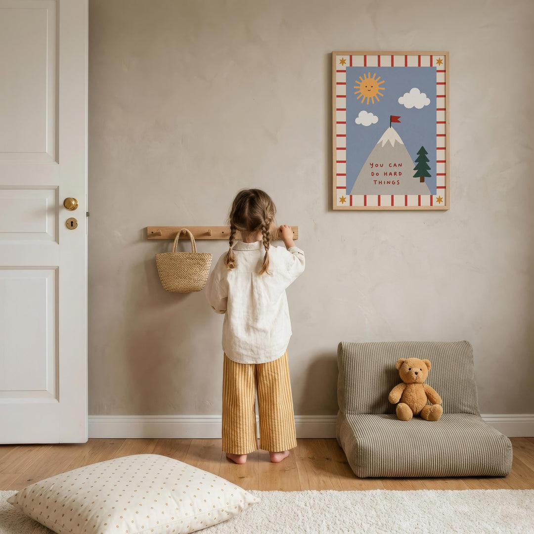 A young girl stands in a cozy room, looking up at a framed picture on the wall. The room has a wooden floor, a chair, and a teddy bear on the floor.