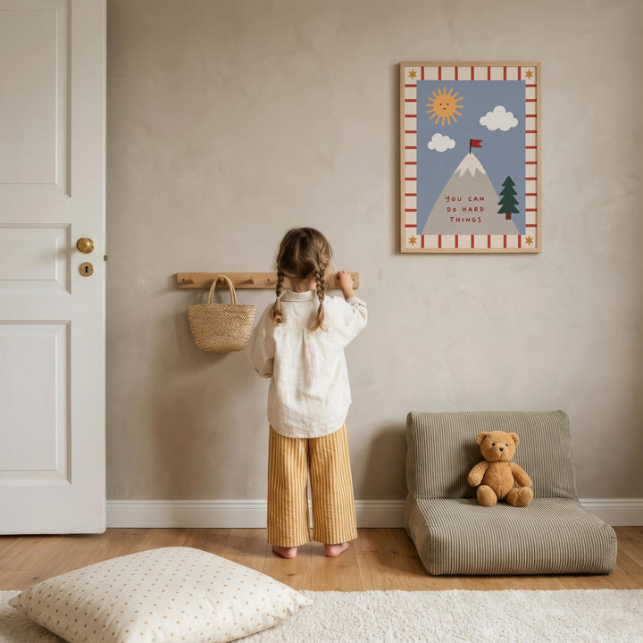 A young girl stands in a cozy room, looking up at a framed picture on the wall. The room has a wooden floor, a chair, and a teddy bear on the floor.