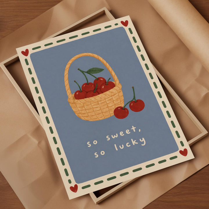 A greeting card with a basket of cherries and the text "So sweet, so lucky" on a blue background.