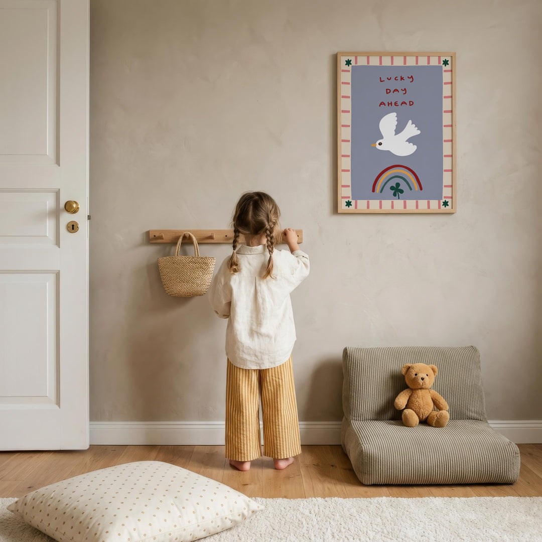 A young girl stands in a cozy room, looking at a framed poster on the wall with a teddy bear on a chair nearby.
