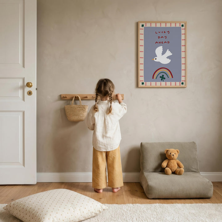 A young girl stands in a cozy room, looking at a framed poster on the wall with a teddy bear on a chair nearby.