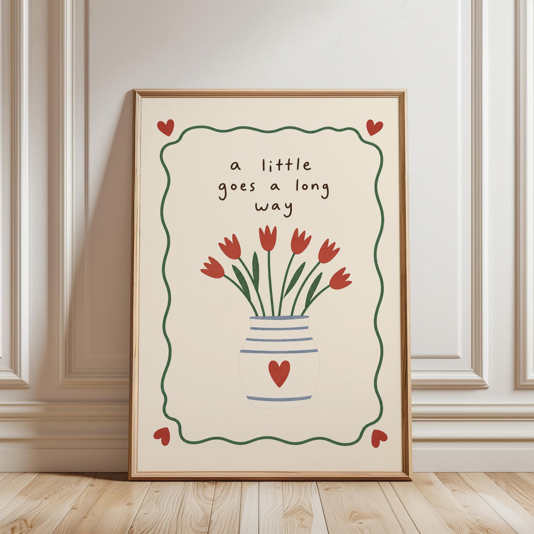 A framed poster with a floral design and the text "a little goes a long way" is displayed on a wooden floor against a white wall.