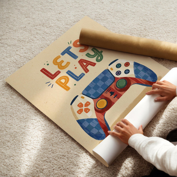 A person's hands are holding a roll of paper, which appears to be a poster or artwork featuring a colorful game controller and the text "LET'S PLAY" written on it.