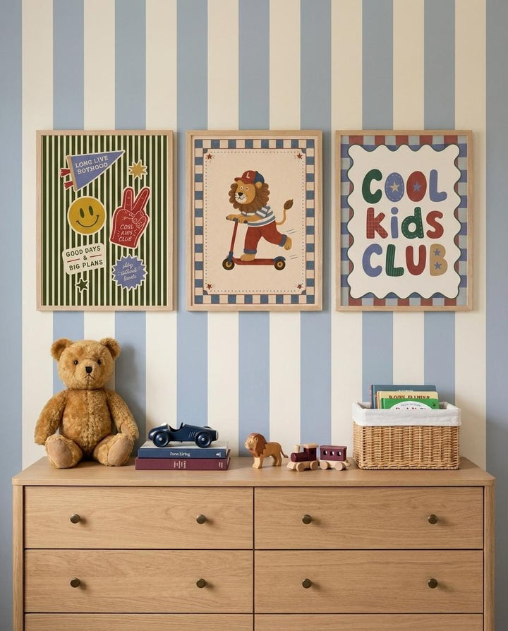 a wooden dresser with three framed pictures on the wall above it. On the dresser, there is a teddy bear, a toy car, and a basket of toys. The wall behind the dresser has blue and white striped wallpaper.
