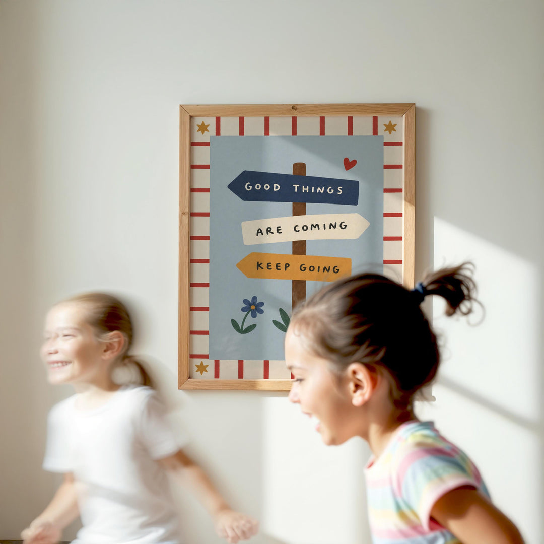 Two young girls are running in front of a framed sign with the text "GOOD THINGS ARE COMING KEEP GOING" and a flower design.