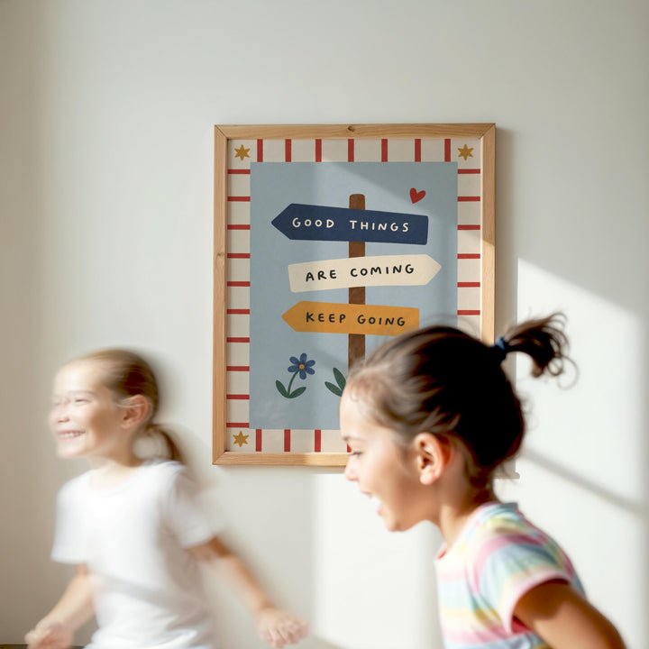Two young girls are running in front of a framed sign with the text "GOOD THINGS ARE COMING KEEP GOING" and a flower design.