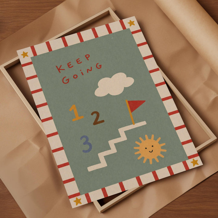 The image is a colorful card with a playful design, featuring a cloud, a flag, and a sun. The card has a message that says "Keep Going" and includes numbers 1, 2, and 3. The card is placed on a brown paper, and there is a wooden frame visible in the background.