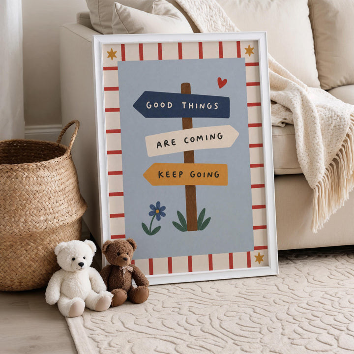 A framed poster with the text "Good Things Are Coming Keep Going" is displayed on a beige couch, accompanied by two teddy bears sitting on the floor.